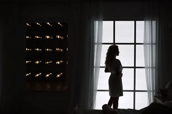 Silhouette of a fashion woman by the window. Wonderful portrait of a girl in a beautiful silk dressing gown. — Stock Photo, Image