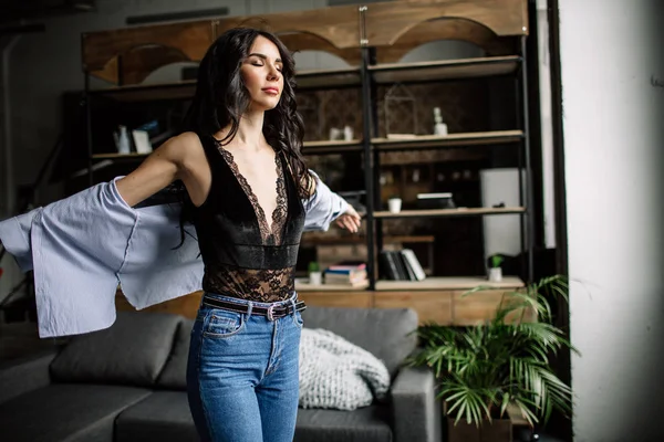 En primer plano. Hermosa morena vestida con una camisa azul y vaqueros en una habitación en el fondo trasero de una estantería y un sofá, estilo de moda —  Fotos de Stock