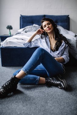 Güzel bir esmer kulaklık kanepe yakınındaki bir odada müzik dinler, bir gömlek ve mavi jeans, mavi giymiş