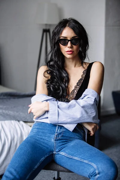 Retrato de uma bela morena em óculos de sol, camisa azul, jeans azul e interior no quarto — Fotografia de Stock