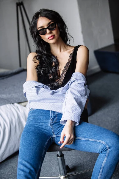 Porträtt av en vacker brunett i solglasögon, blå tröja, blå jeans och inredning i rum — Stockfoto