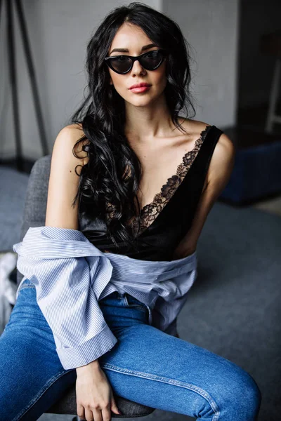 Retrato de una hermosa morena en gafas de sol, camisa azul, vaqueros azules e interior en la habitación — Foto de Stock