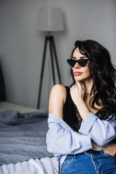 Retrato de una hermosa morena en gafas de sol, camisa azul, vaqueros azules e interior en la habitación —  Fotos de Stock