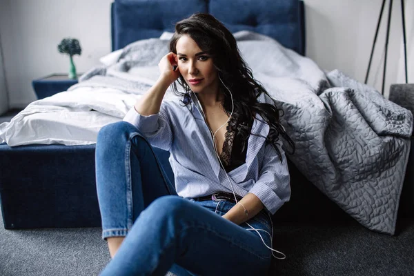 Piękna Brunetka słucha muzyki w słuchawkach w pokoju w pobliżu kanapie, jest ubrana w koszulę i niebieskie dżinsy, niebieski — Zdjęcie stockowe
