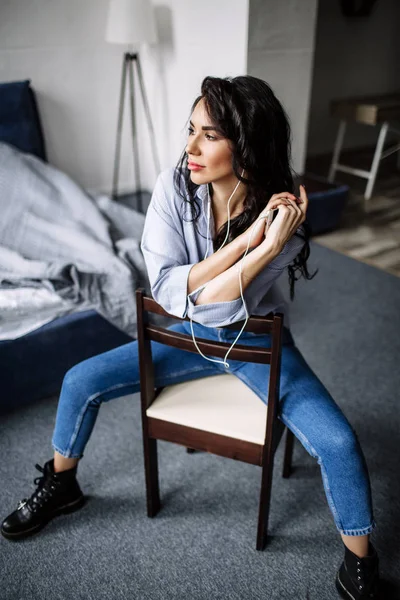 Una hermosa morena escucha música en auriculares en una habitación cerca del sofá, está vestida con una camisa y vaqueros azules, azul — Foto de Stock