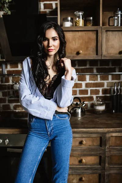 Belle brune dans la cuisine, femme au foyer moderne, mode, mobilier marron en bois, vêtue d'un jean bleu et d'une chemise bleue — Photo