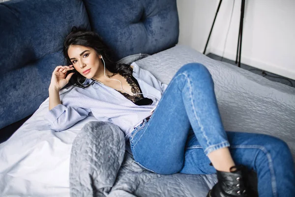 En vacker brunett lyssnar på musik i hörlurar i ett rum nära soffan, är klädd i en tröja och blå jeans, blå — Stockfoto