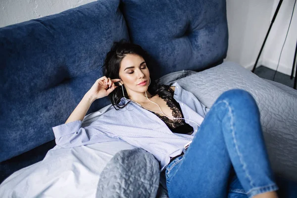 Μια όμορφη μελαχρινή ακούει μουσική με ακουστικά σε ένα δωμάτιο κοντά στο καναπέ, είναι ντυμένος με ένα πουκάμισο και μπλε τζιν, μπλε — Φωτογραφία Αρχείου