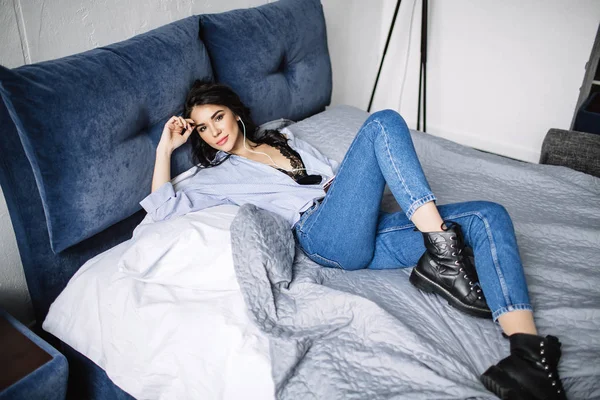 En vacker brunett lyssnar på musik i hörlurar i ett rum nära soffan, är klädd i en tröja och blå jeans, blå — Stockfoto