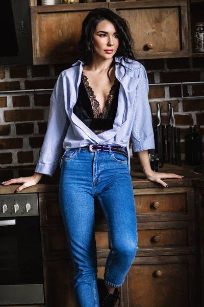 Belle brune dans la cuisine, femme au foyer moderne, mode, mobilier marron en bois, vêtue d'un jean bleu et d'une chemise bleue — Photo