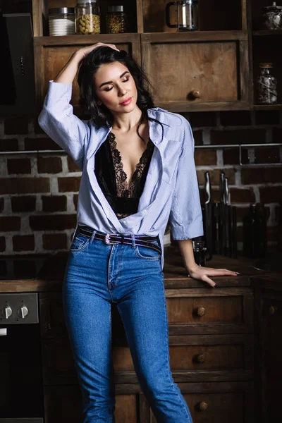 Vacker brunett i kök, modern hemmafru, mode, bruna trämöbler, klädd i blå jeans och en blå skjorta — Stockfoto