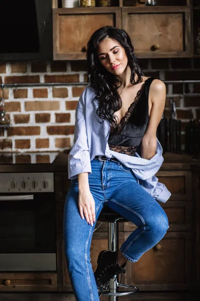 Linda morena na cozinha, dona de casa moderna, moda, móveis castanhos de madeira, vestidos com jeans azul e uma camisa azul — Fotografia de Stock