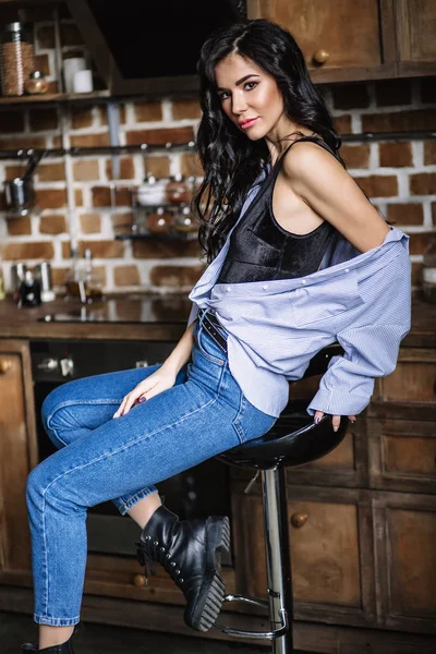 Mooie brunette in de keuken, moderne huisvrouw, fashion, houten bruin meubilair, gekleed in blauwe spijkerbroek en een blauw shirt — Stockfoto