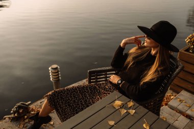 Çalışan genç bir iş kadını. Kafasına siyah bir şapka ve güneş gözlüğü takıyor. Yatın arka planı, su.
