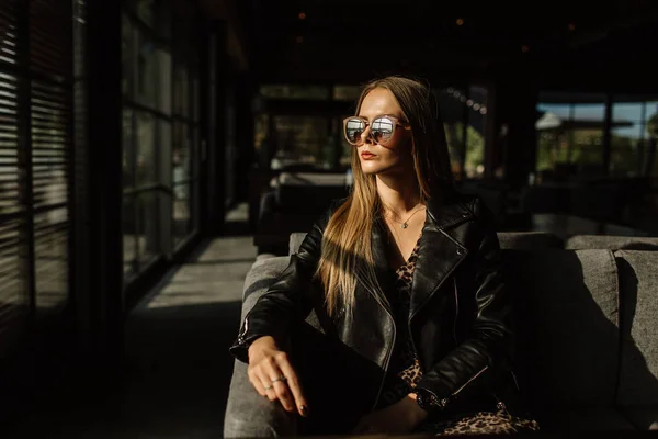 Belle fille modèle dans un restaurant dans un bel intérieur loft. sur les yeux des lunettes de soleil, vêtus d'une veste noire en cuir, vêtements décontractés. photographie chaude, lumière du soleil — Photo