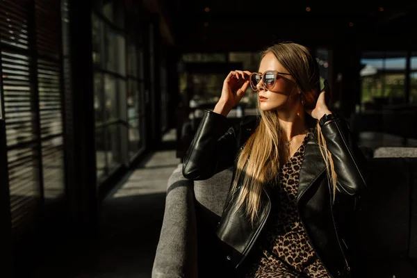 Belle fille modèle dans un restaurant dans un bel intérieur loft. sur les yeux des lunettes de soleil, vêtus d'une veste noire en cuir, vêtements décontractés. photographie chaude, lumière du soleil — Photo
