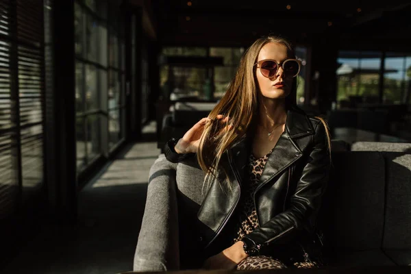 Belle fille modèle dans un restaurant dans un bel intérieur loft. sur les yeux des lunettes de soleil, vêtus d'une veste noire en cuir, vêtements décontractés. photographie chaude, lumière du soleil — Photo