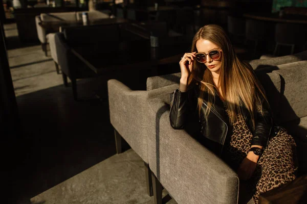 Красивая девушка-модель в ресторане в красивом интерьере лофта. на глазах солнцезащитных очков, одет в кожаную черную куртку, повседневную одежду. теплое фотографирование, солнечный свет — стоковое фото