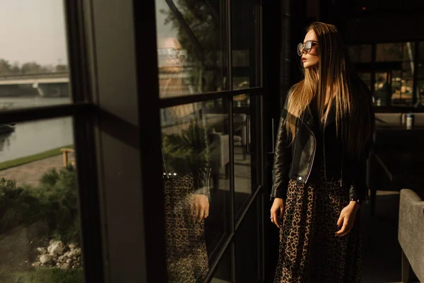 Modelo hermosa chica en un restaurante en un hermoso loft interior. en los ojos de las gafas de sol, vestido con una chaqueta de cuero negro, ropa casual. fotografía cálida, luz solar — Foto de Stock