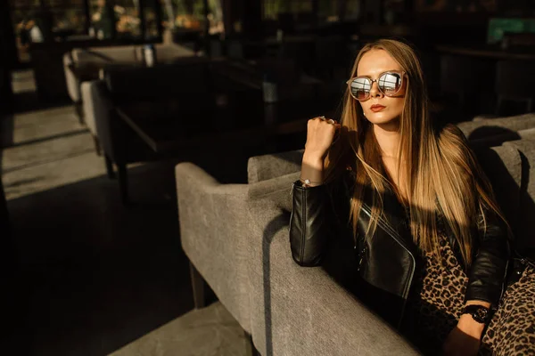 Όμορφο κορίτσι μοντέλο σε ένα εστιατόριο σε ένα όμορφο εσωτερικό πατάρι. στα μάτια των γυαλιών ηλίου, ντυμένοι με ένα μαύρο δερμάτινο μπουφάν, casual ρούχα. ζεστή φωτογραφία, φως του ήλιου — Φωτογραφία Αρχείου