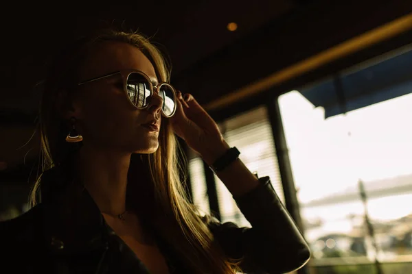 Modelo hermosa chica en un restaurante en un hermoso loft interior. en los ojos de las gafas de sol, vestido con una chaqueta de cuero negro, ropa casual. fotografía cálida, luz solar — Foto de Stock