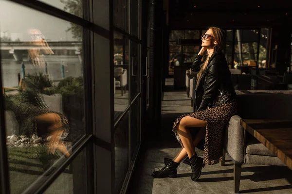 Schöne Mädchen Modell in einem Restaurant in einem schönen Loft-Interieur. Sonnenbrille auf, schwarze Lederjacke, lässige Kleidung. warme Fotografie, Sonnenlicht — Stockfoto