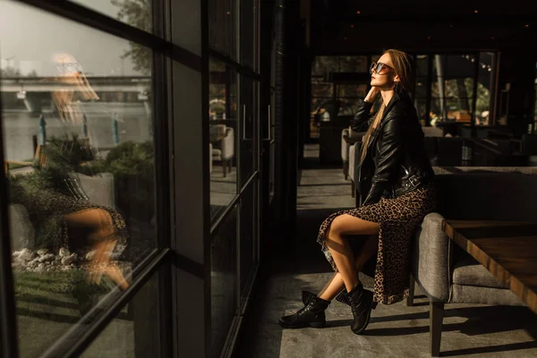 Modelo hermosa chica en un restaurante en un hermoso loft interior. en los ojos de las gafas de sol, vestido con una chaqueta de cuero negro, ropa casual. fotografía cálida, luz solar — Foto de Stock