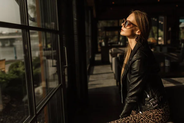 Modelo hermosa chica en un restaurante en un hermoso loft interior. en los ojos de las gafas de sol, vestido con una chaqueta de cuero negro, ropa casual. fotografía cálida, luz solar — Foto de Stock