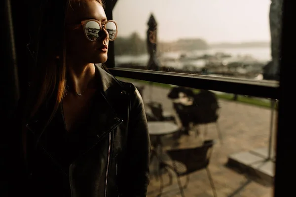 Modelo hermosa chica en un restaurante en un hermoso loft interior. en los ojos de las gafas de sol, vestido con una chaqueta de cuero negro, ropa casual. fotografía cálida, luz solar —  Fotos de Stock