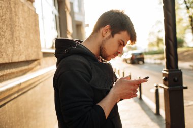 Mutlu gülümseyen genç adam şehir merkezinde gün batımında yaya geçidinde dururken modern akıllı telefonu kullanıyor.