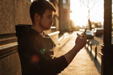 Mutlu gülümseyen genç adam şehir merkezinde gün batımında yaya geçidinde dururken modern akıllı telefonu kullanıyor.