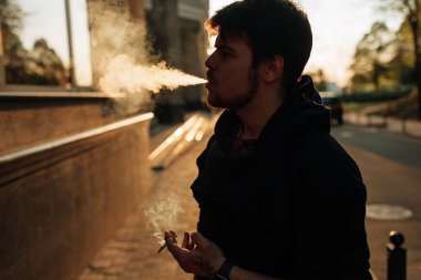 Genç adam şehrin ortasında gün batımında sigara içiyor.