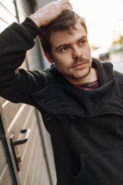 Açık havada duran ve duvara yaslanan şık, yakışıklı genç bir adamın portresi. Siyah kapüşonlu adam