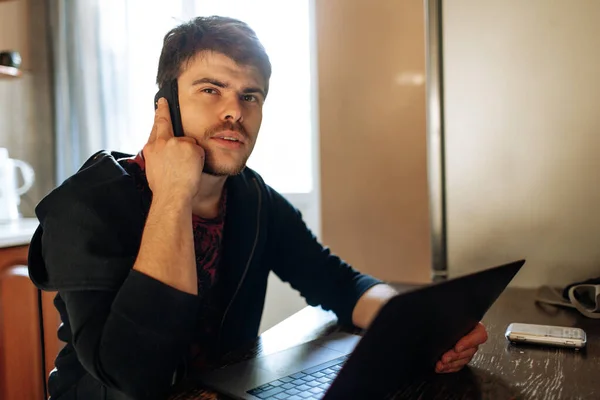 一位留着胡子的年轻嬉皮士身穿黑色夹克 坐在厨房桌子旁 用笔记本电脑 用手机通话 网络营销 电子学习 启动阶段 — 图库照片