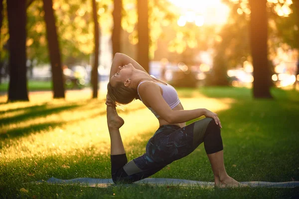 Belle jeune femme pratique le yoga asana Roi Pigeon pose rajakapotasana dans le parc au coucher du soleil — Photo