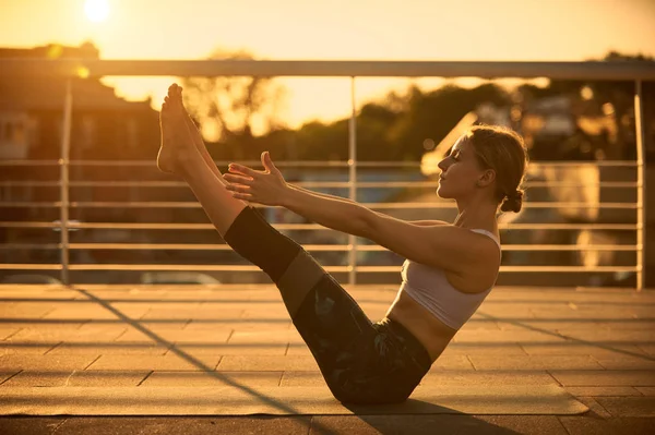 Молодая женщина практикующая йогу, сидящая в Paripurna Navasana упражнения на террасе на закате — стоковое фото