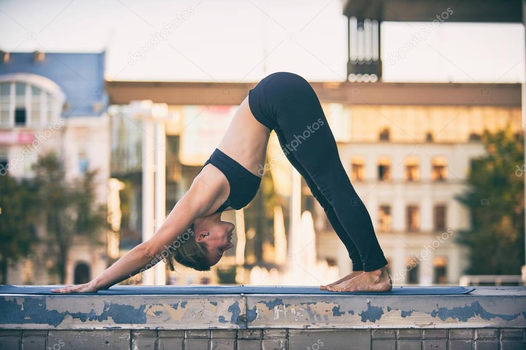 Beautiful young woman practices yoga asana downward facing dog in the city