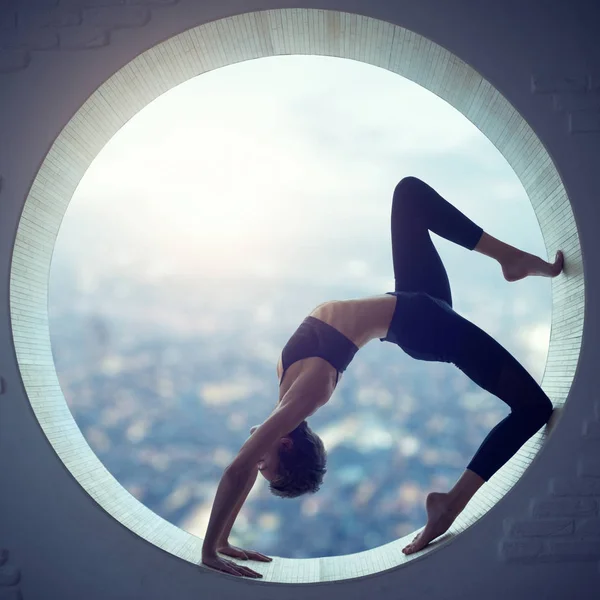 Beautiful sporty fit yogi woman practices yoga asana Eka Pada Urdhva Dhanurasana in a round window with a view of the city — Stock Photo, Image