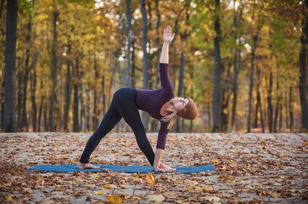 美丽的年轻女子练习瑜伽体式三角姿势三角在秋天公园的木甲板上 — 图库照片