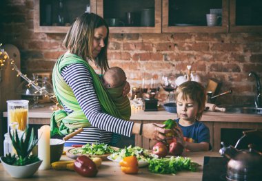 Mutlu genç bir aile, iki çocuklu güzel bir anne, sevimli bir anaokulu çocuğu ve bebek, güneşli bir mutfakta birlikte yemek pişiriyorlar..
