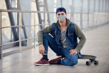Kapüşonlu yakışıklı beyaz bir adam sokakta kaykayın üzerinde oturuyor ve yüzünde tıbbi maske var. Coronavirus konsept fotoğrafı.