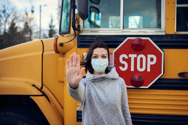 A woman in a protective mask stands on the background of a school bus. Raises a hand and stops the epidemic of coronavirus. A large stop sign is visible on the background