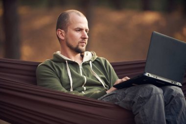 Man Hiker, çam ormanında hamakta dizüstü bilgisayarla çalışıyor. Uzak çalışma konsepti.