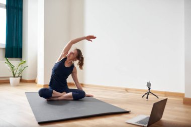 İnternette yoga yapan bir kadın. Oturma odasında bilgisayar ve kamera var. Sağlıklı yaşam tarzı ve mesafe çalışma kavramları.