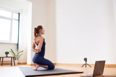 İnternette yoga yapan bir kadın. Oturma odasında bilgisayar ve kamera var. Sağlıklı yaşam tarzı ve mesafe çalışma kavramları.