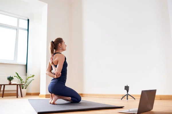 Woman doing yoga online. Computer and camera in her living room. Healthy lifestyle and distance work concepts.