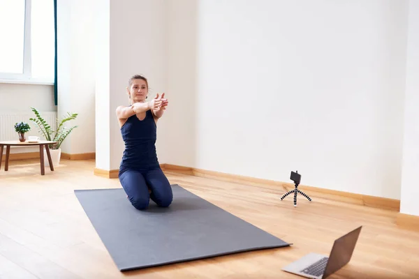 Woman doing yoga online. Computer and camera in her living room. Healthy lifestyle and distance work concepts.