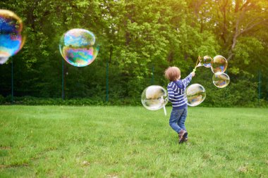 Çocuk parkındaki yeşil çimlere sabun köpüğü üfleyen sevimli anaokulu çocuğunun açık hava portresi.