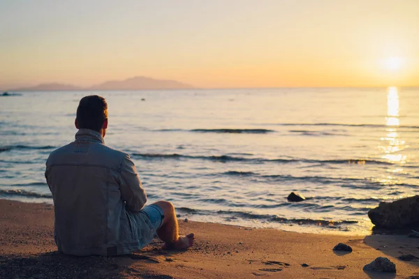 Genç bir adam deniz sahilde oturan ve güneş doğarken arıyorsunuz