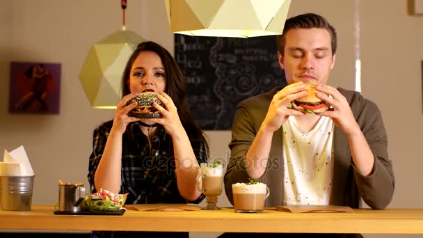 Fiatal nő, és a fiatalember harap hamburgert a burger bárban — Stock videók